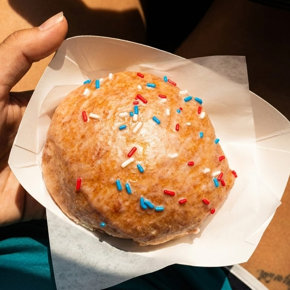 A hand holds a glazed donut with red, white, and blue sprinkles in a paper tray.